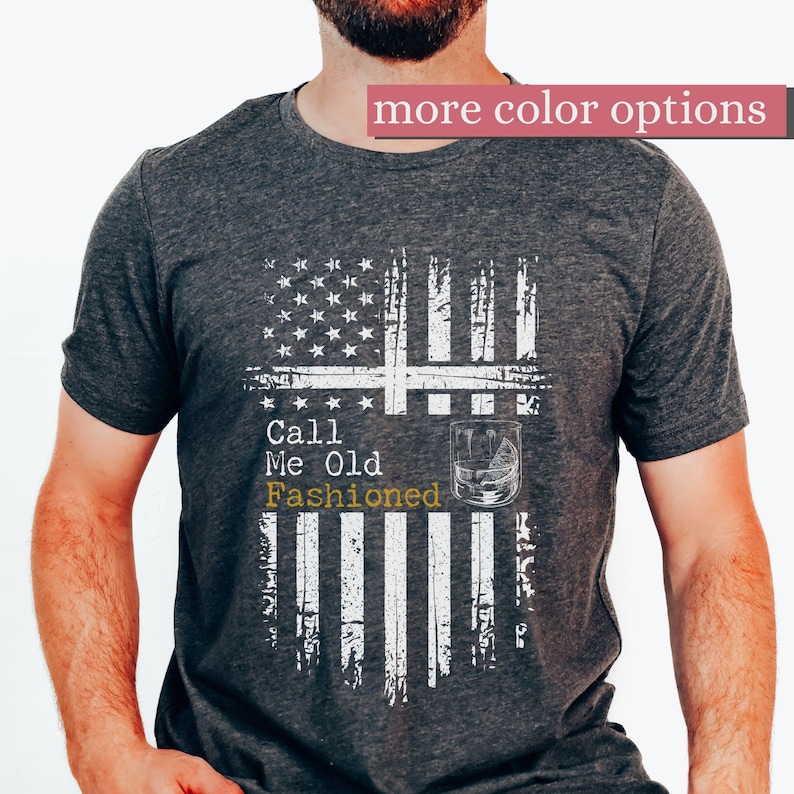 May include: A dark heather gray short-sleeve t-shirt featuring a distressed white graphic of the American flag. The design includes the text Call Me Old Fashioned and an illustration of a glass of whiskey with ice on the front of the shirt.
