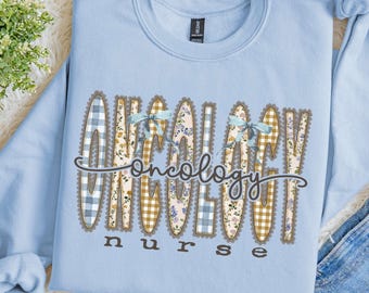 Puede incluir: Una sudadera de cuello redondo azul claro con la palabra ONCOLOGY en letras grandes estampadas con borde festoneado. Las letras tienen cuadros vichy azules, flores amarillas y cuadros marrones. La palabra nurse aparece debajo.