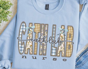 Puede incluir: Una sudadera de cuello redondo azul claro con el texto "cath lab nurse" en una fuente decorativa. Las letras están rellenas con patrones de cuadros vichy azules y blancos, amarillos y blancos, y motivos florales, con dos lazos azul claro.