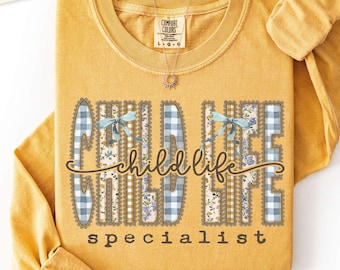 Puede incluir: Una camiseta de manga larga color amarillo mostaza con cuello redondo. El frente presenta el texto "child life specialist" en letras marrones cursivas y de molde, decorado con patrones de cuadros azul claro, flores y lazos azules.