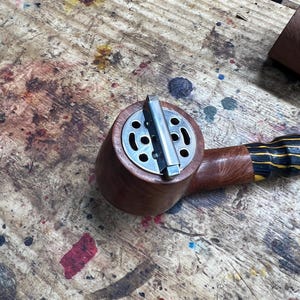 May include: A wooden tobacco pipe with a metal top and a striped handle. The pipe is brown with a silver metal top and a black and yellow striped handle. The pipe is on a wooden surface.
