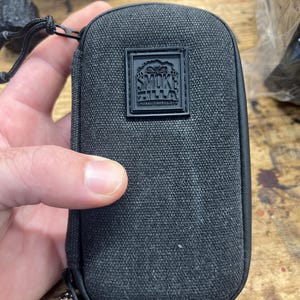 Puede incluir: Bolsa negra con cremallera y el texto "SMOKE ZILLA" en un parche cuadrado. La bolsa se sostiene en una mano y tiene un pequeño lazo con un cierre de metal.