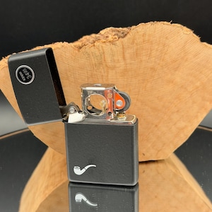 May include: Black Zippo lighter with a pipe logo, open to reveal the lighter's interior. The lighter has a silver hinge and a small orange wheel. The background features a wooden surface.