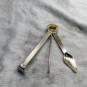 Puede incluir: Una herramienta de pipa de metal plateado con una cuchara, un atizador y un escariador. El mango tiene un diseño de llave griega. La cuchara está a la derecha, el atizador en el medio y el escariador a la izquierda.