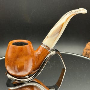 May include: A handcrafted tobacco pipe with a polished, light brown bowl and a cream-colored, curved stem. The pipe rests on a silver-colored stand, showcasing the wood grain and intricate design. The stem features a decorative band.
