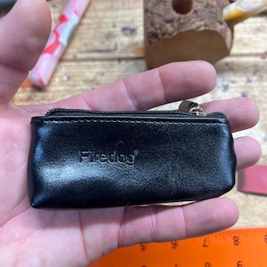 Puede incluir: Pequeño estuche de cuero negro con cierre de cremallera. La palabra "Firedog" está impresa en la parte delantera. El estuche es rectangular y parece estar vacío. Una regla está en el fondo.