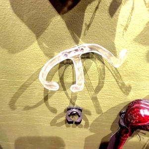 May include: Clear plastic bicycle handlebar with curved ends and a small black ring. The handlebar is positioned above the ring, with a red object partially visible on the right side of the image.