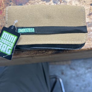 May include: A tan canvas mini tobacco bag with a black elastic band and the text "SMOKEZILLA". A green and white tag reads "MINI TOBACCO BAG". The bag is rectangular and appears to be made of durable material.