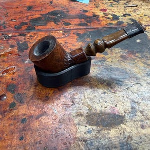 Könnte beinhalten: Eine handgefertigte Tabakpfeife mit einer dunkelbraunen Pfeifenkopf und einem helleren braunen Mundstück. Die Pfeife ruht auf einem schwarzen Ständer, der ihr aufwendiges Design zeigt. Die Holzmaserung ist sichtbar und trägt zu ihrem rustikalen Charme bei.