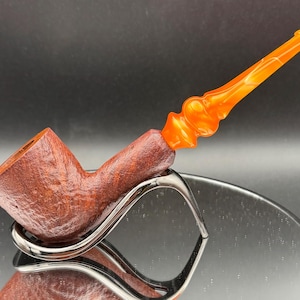 May include: A handcrafted tobacco pipe with a brown bowl and an orange, swirled stem. The pipe is displayed on a silver stand against a dark background. The bowl has a textured finish.