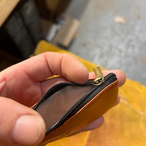 Peut inclure: Petite pochette en cuir orange ouverte, avec une fermeture &eacute;clair noire et un curseur dor&eacute;. L'int&eacute;rieur de la pochette est gris clair. La pochette est tenue ouverte, r&eacute;v&eacute;lant l'int&eacute;rieur.