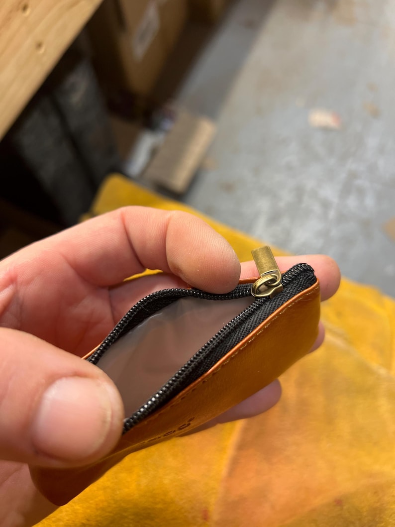 Peut inclure: Petite pochette en cuir orange ouverte, avec une fermeture &eacute;clair noire et un curseur dor&eacute;. L'int&eacute;rieur de la pochette est gris clair. La pochette est tenue ouverte, r&eacute;v&eacute;lant l'int&eacute;rieur.