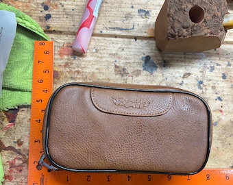Puede incluir: Un estuche de cuero marrón con la palabra "Scotte" en relieve en la solapa. El estuche es rectangular con un ribete negro y un cierre de cremallera. Una regla muestra que el estuche mide aproximadamente 23 cm de largo. Varias herramientas de carpintería están en el fondo.