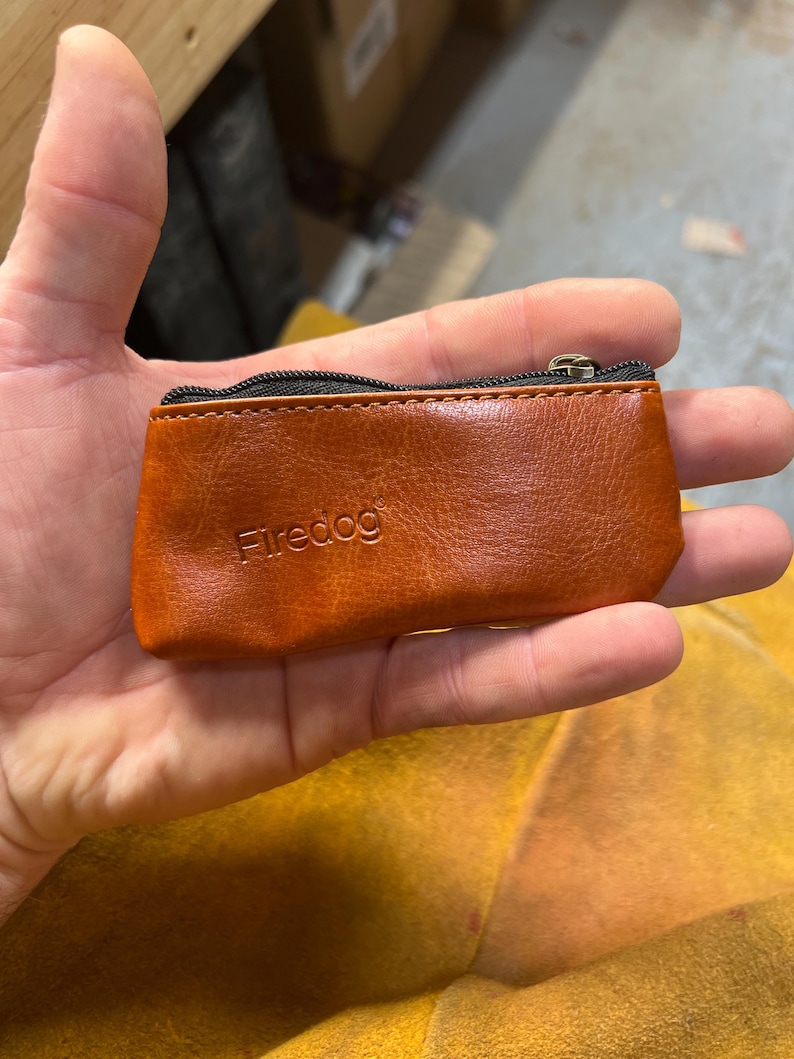 Peut inclure: Un petit porte-monnaie rectangulaire en cuir marron avec une fermeture &eacute;clair noire et le mot "Firedog" en relief. Le porte-monnaie est tenu dans une main et semble neuf.