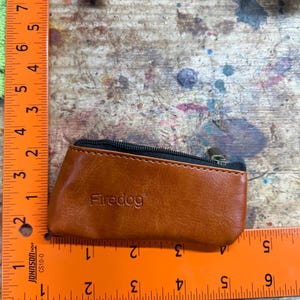 Peut inclure: Un petit porte-monnaie en cuir marron avec une fermeture &eacute;clair noire et le mot "Firedog" en relief sur le devant. Le porte-monnaie mesure environ 10 cm de long. Le fond est une surface en bois avec des &eacute;claboussures de peinture.
