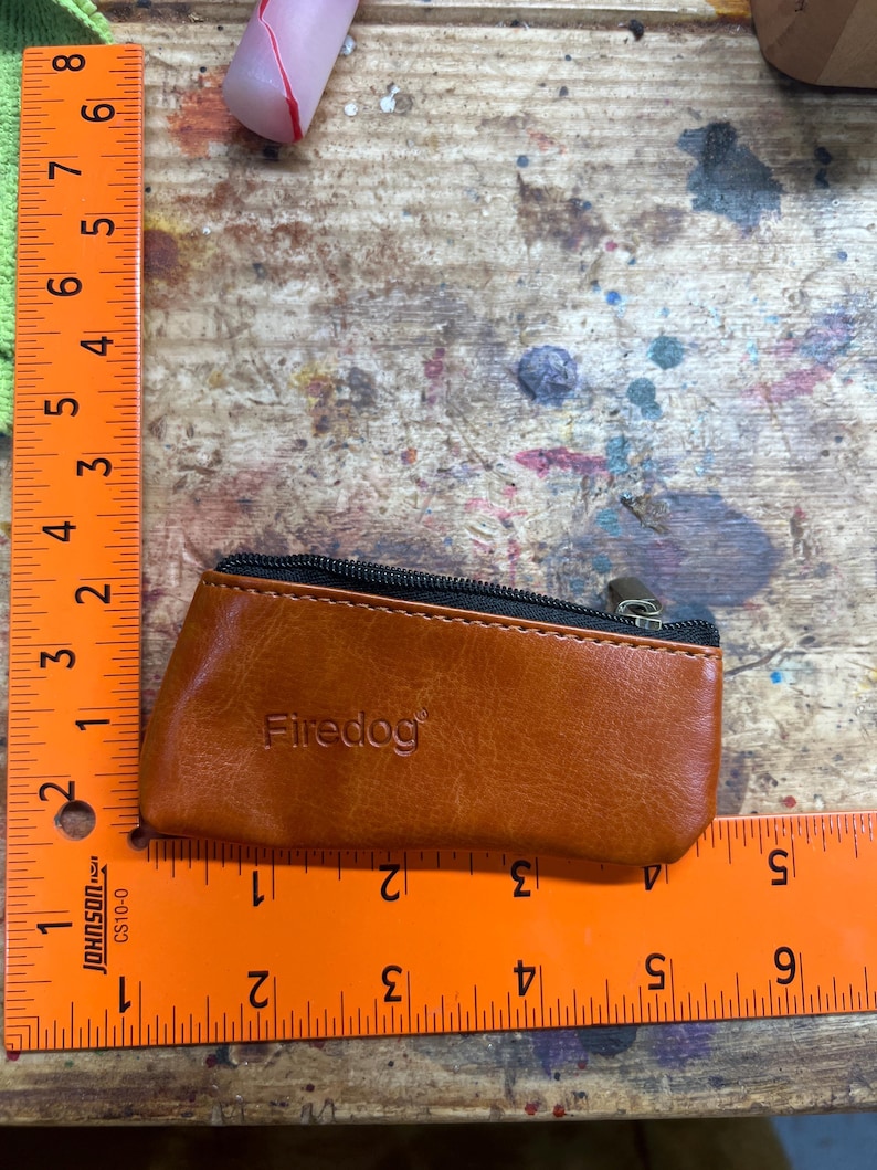 Peut inclure: Un petit porte-monnaie en cuir marron avec une fermeture &eacute;clair noire et le mot "Firedog" en relief sur le devant. Le porte-monnaie mesure environ 10 cm de long. Le fond est une surface en bois avec des &eacute;claboussures de peinture.
