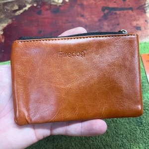Puede incluir: Un monedero rectangular de cuero marrón con cierre de cremallera negro. La palabra "Firedog" está grabada en la parte delantera. El monedero está en una mano.