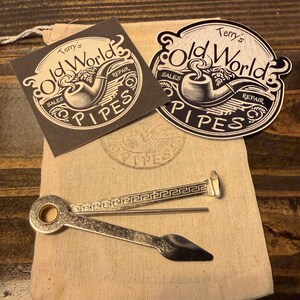 May include: A silver-toned metal pipe tool with a textured handle and a flat, pointed end, resting on a beige fabric pouch. Two stickers are included, each featuring the text Terry's Old World PIPES with a graphic illustration of a smoking pipe.