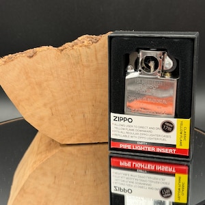 May include: A Zippo pipe lighter insert in a black box, with the Zippo logo and product details visible. The insert is silver-toned, and the box has a yellow and red label. The background includes a wooden element and a reflective surface.