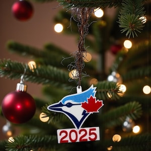 Puede incluir: Adorno navideño con el logo de los Toronto Blue Jays, una hoja de arce roja y el año 2025. El adorno blanco, azul y rojo cuelga de un árbol de Navidad con bolas rojas y plateadas.