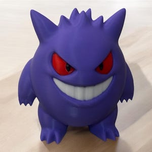 Könnte beinhalten: Eine lila Gengar-Figur mit roten Augen und einem breiten Grinsen. Die Figur hat stachelige Vorsprünge auf dem Kopf und steht. Diese Sammlerfigur ist ein beliebtes Charakter.