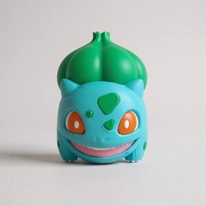Op de afbeelding: Een Bulbasaur-beeldje in een staande pose. Het beeldje is voornamelijk blauwgroen met een groene bol op de rug. Het heeft oranje ogen, een lachende mond en groene vlekken. Verzamelaarsitem.