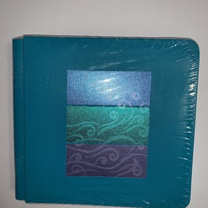 May include: A teal photo album wrapped in clear plastic. The front cover features a rectangular design with three horizontal sections: blue, teal, and purple, each with a swirling pattern. The album is rectangular and appears new.