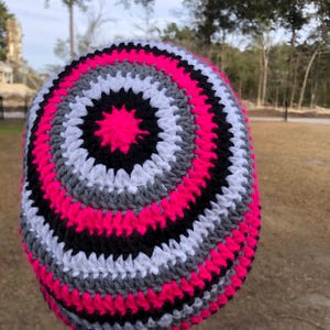 May include: A handmade, crocheted beanie hat with a vibrant, concentric design. The hat features alternating bands of bright pink, black, white, and gray yarn. The hat is a stylish accessory for cold weather.