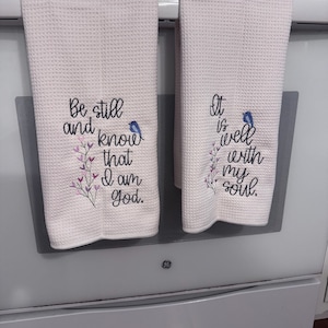 Op de afbeelding: Twee lichtroze keukenhanddoeken met wafelweefsel. Elke handdoek heeft geborduurde tekst en een kleine blauwe vogel. Op de ene handdoek staat "Be still and know that I am God." De andere luidt "It is well with my soul."
