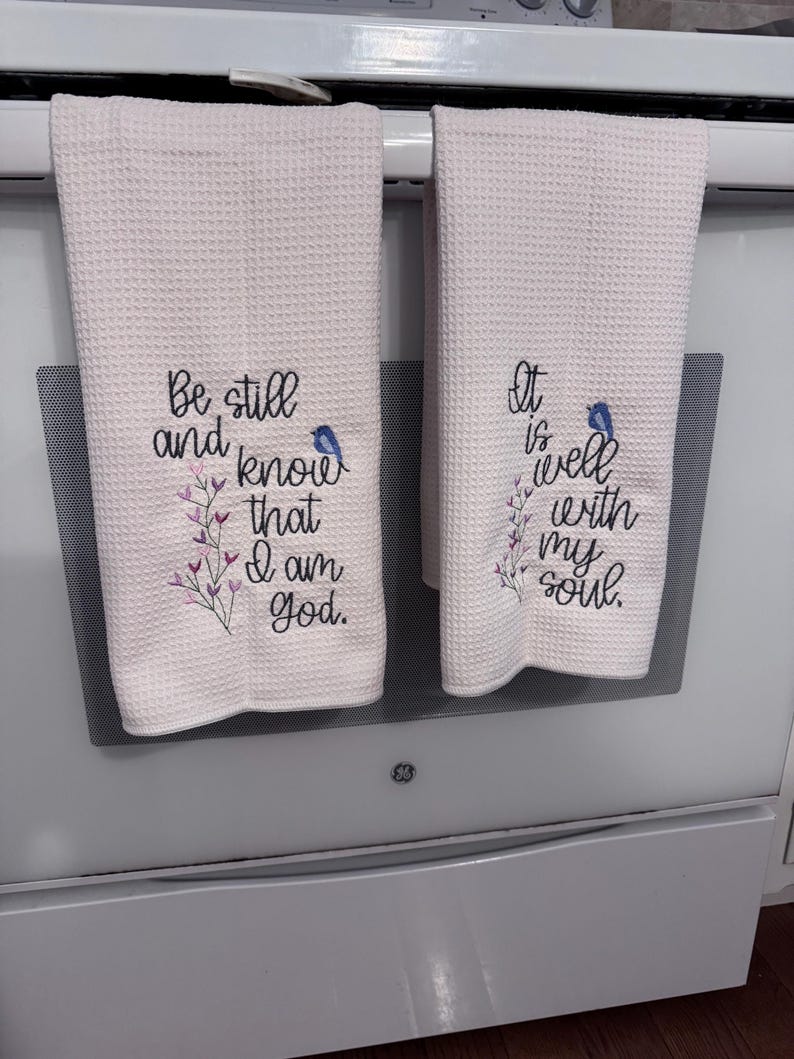 Op de afbeelding: Twee lichtroze keukenhanddoeken met wafelweefsel. Elke handdoek heeft geborduurde tekst en een kleine blauwe vogel. Op de ene handdoek staat "Be still and know that I am God." De andere luidt "It is well with my soul."