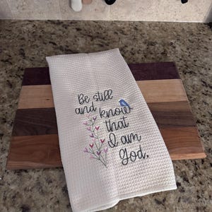 Op de afbeelding: Witte keukenhanddoek met wafelweefsel en geborduurde tekst "Be still and know that I am God." Voorzien van een bloemmotief en een kleine blauwe vogel. De handdoek ligt op een houten snijplank met diverse houttinten.