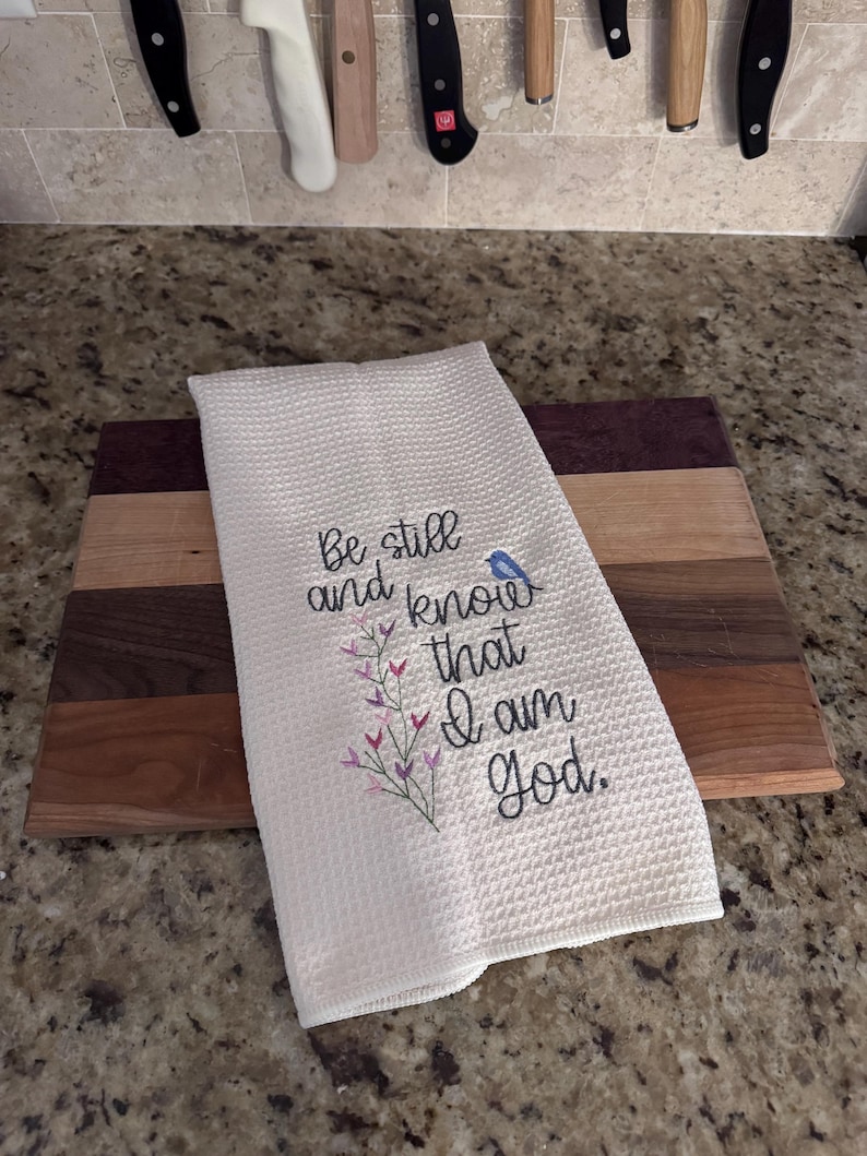 Op de afbeelding: Witte keukenhanddoek met wafelweefsel en geborduurde tekst "Be still and know that I am God." Voorzien van een bloemmotief en een kleine blauwe vogel. De handdoek ligt op een houten snijplank met diverse houttinten.
