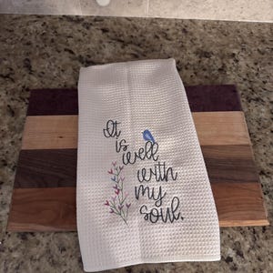 Op de afbeelding: Witte keukenhanddoek met wafelweefsel en geborduurde tekst "It is well with my soul" en een kleine blauwe vogel. De handdoek ligt op een houten snijplank met bruine en paarse strepen.