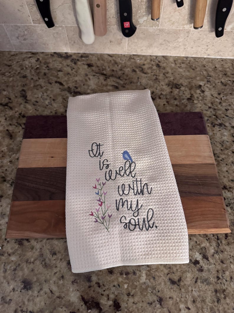 Op de afbeelding: Witte keukenhanddoek met wafelweefsel en geborduurde tekst "It is well with my soul" en een kleine blauwe vogel. De handdoek ligt op een houten snijplank met bruine en paarse strepen.