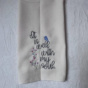 Op de afbeelding: Cr&egrave;mekleurige keukentowel met wafelweefsel geborduurd met de zin "It is well with my soul". Een kleine blauwe vogel en paarse bloemaccenten zijn geborduurd bij de tekst. De handdoek is dubbelgevouwen.