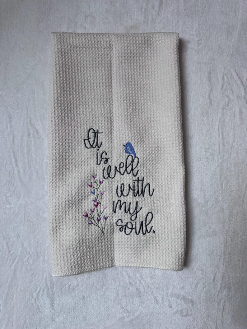 Op de afbeelding: Cr&egrave;mekleurige keukentowel met wafelweefsel geborduurd met de zin "It is well with my soul". Een kleine blauwe vogel en paarse bloemaccenten zijn geborduurd bij de tekst. De handdoek is dubbelgevouwen.