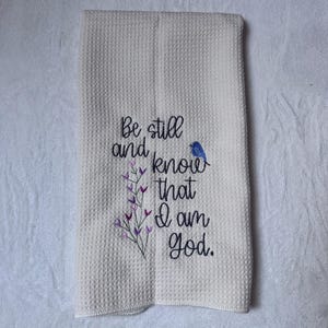 Op de afbeelding: Cr&egrave;mekleurige wafelgeweven keukendoek met geborduurde tekst en bloemaccenten. De tekst luidt "Be still and know that I am God." Een kleine blauwe vogel is naast de tekst geborduurd. Paarse en groene bloemenborduursels zijn ook aanwezig.