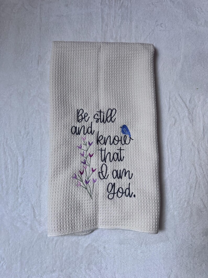 Op de afbeelding: Cr&egrave;mekleurige wafelgeweven keukendoek met geborduurde tekst en bloemaccenten. De tekst luidt "Be still and know that I am God." Een kleine blauwe vogel is naast de tekst geborduurd. Paarse en groene bloemenborduursels zijn ook aanwezig.