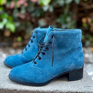 May include: A pair of ankle boots in a vibrant blue suede material featuring a rounded toe, a lace-up front with black laces, and a low black block heel. The boots have a smooth, consistent texture and a clean, simple design with visible stitching.