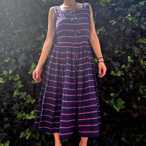 May include: A sleeveless midi dress in a deep navy blue featuring horizontal stripes in white and red. The bodice and skirt are connected at the waist, with a vertical row of small decorative bows running down the center front of the garment.