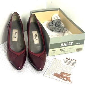 Può includere: Un paio di décolleté Bally in pelle bordeaux con punta affilata e rifiniture rosse a contrasto. Le scarpe sono esposte accanto alla loro scatola originale e a una guida per la cura. La scatola è etichettata "BALLY" e "MADE IN ITALY".