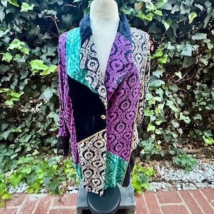 Op de afbeelding: Een blazer met lange mouwen en een patchworkontwerp met panelen van zwart fluweel, groenblauwe stof en een paars met zwart damastpatroon. Het kledingstuk heeft een ingekeepte kraag, een enkele goudkleurige knoopsluiting en een gestructureerd silhouet.