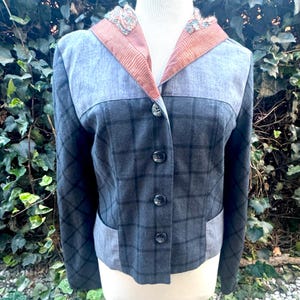 Puede incluir: Un blazer a cuadros gris con un diseño único. La chaqueta presenta una combinación de colores gris y azul con un cuello en contraste y cierre de botones. El blazer tiene un corte entallado y se exhibe en un maniquí.