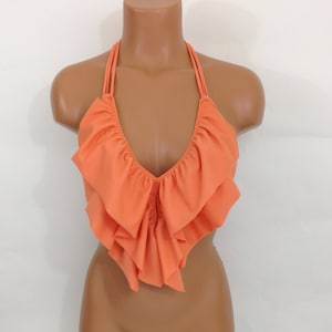 May include: An orange halter top with a ruffled design. The top features a deep V-neckline and thin straps that tie around the neck. The ruffled fabric adds a decorative element to the garment. The top is displayed on a mannequin.