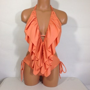 May include: A peach-colored swimsuit with a halter neck and a deep V-neckline. The swimsuit features a ruffled design across the chest and waist, with side cutouts and tie closures. The swimsuit is displayed on a mannequin.