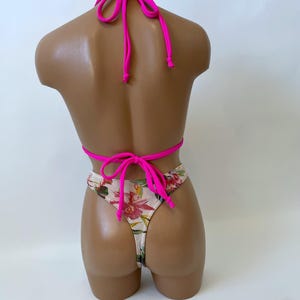 May include: A two-piece swimsuit displayed on a mannequin. The top is a bright pink halter style, and the bottom is a thong with a floral pattern of red, pink, and green flowers on a white background. The swimsuit is tied with pink strings.