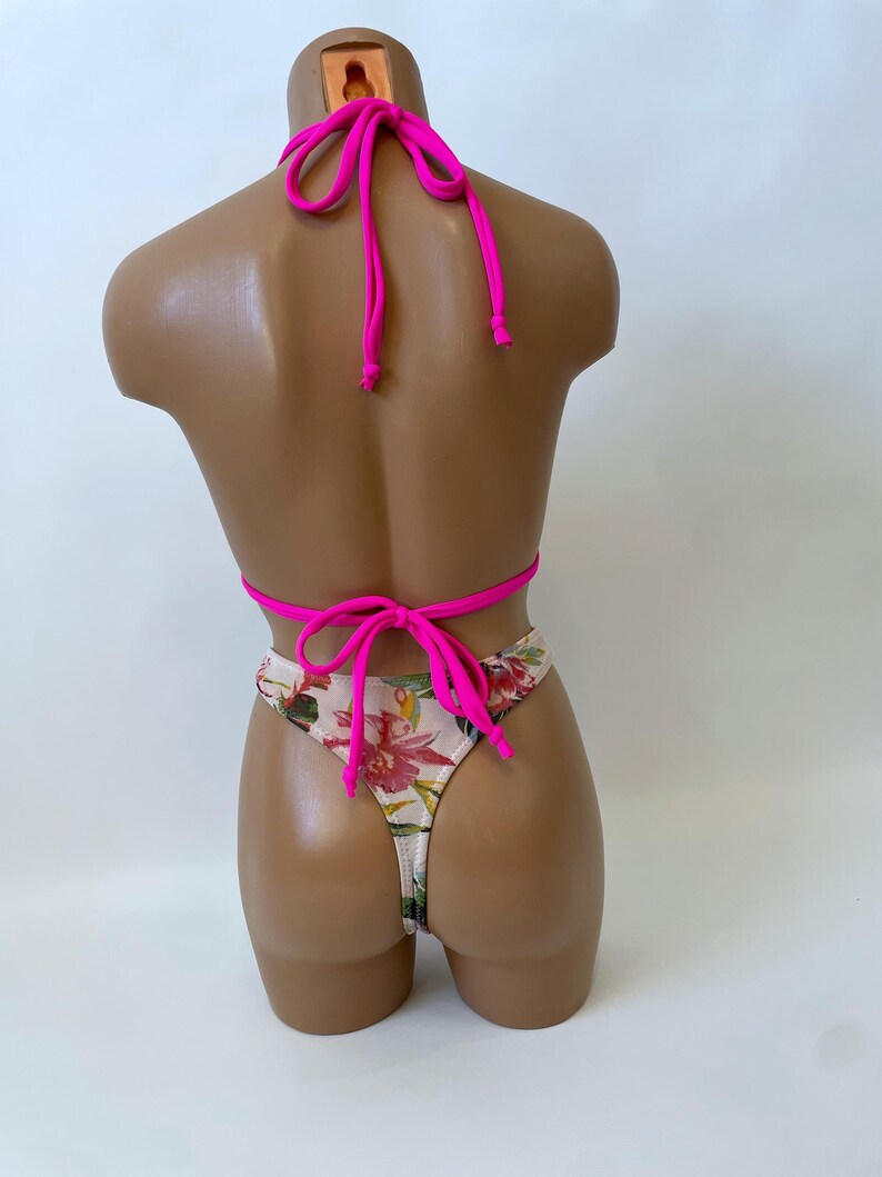 May include: A two-piece swimsuit displayed on a mannequin. The top is a bright pink halter style, and the bottom is a thong with a floral pattern of red, pink, and green flowers on a white background. The swimsuit is tied with pink strings.