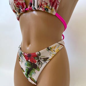 May include: A two-piece bikini set with a floral pattern. The top is a triangle style with hot pink straps. The bottoms are a thong style. The floral pattern includes red, pink, green, and yellow flowers and leaves on a white background.