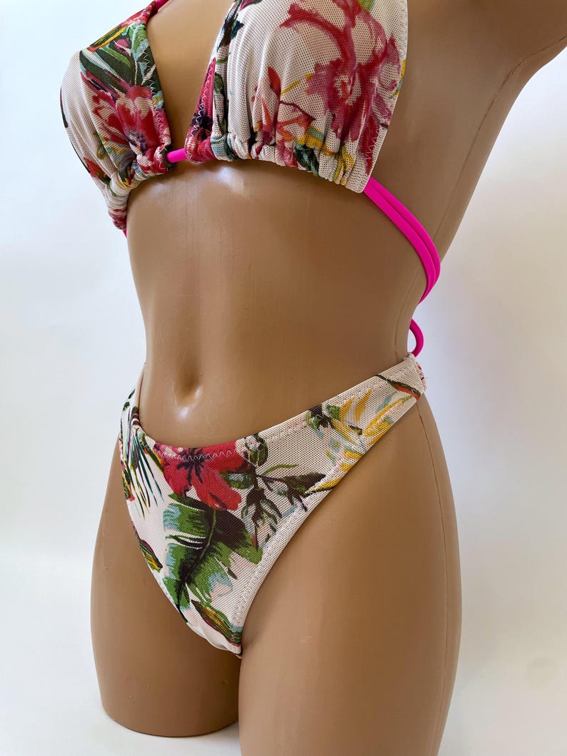 May include: A two-piece bikini set with a floral pattern. The top is a triangle style with hot pink straps. The bottoms are a thong style. The floral pattern includes red, pink, green, and yellow flowers and leaves on a white background.