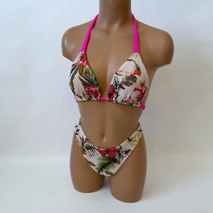May include: A two-piece bikini set with a floral pattern. The bikini top has a halter neck with bright pink straps. The bottoms are high-waisted. The floral pattern includes red, green, and yellow flowers on a cream background.
